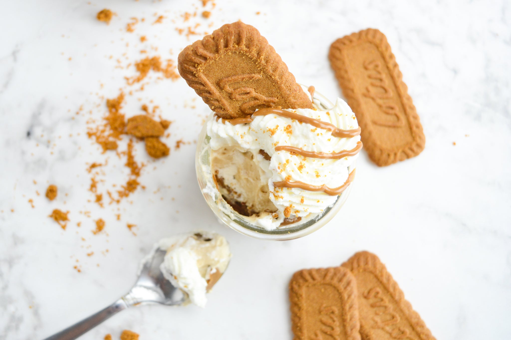 overhead view of marbled table top with cheesecake cup with whipped cream and drizzle of cookie butter a spoon with whipped cream sits to the side and lotus biscoff cookies and crumbs surround the cup