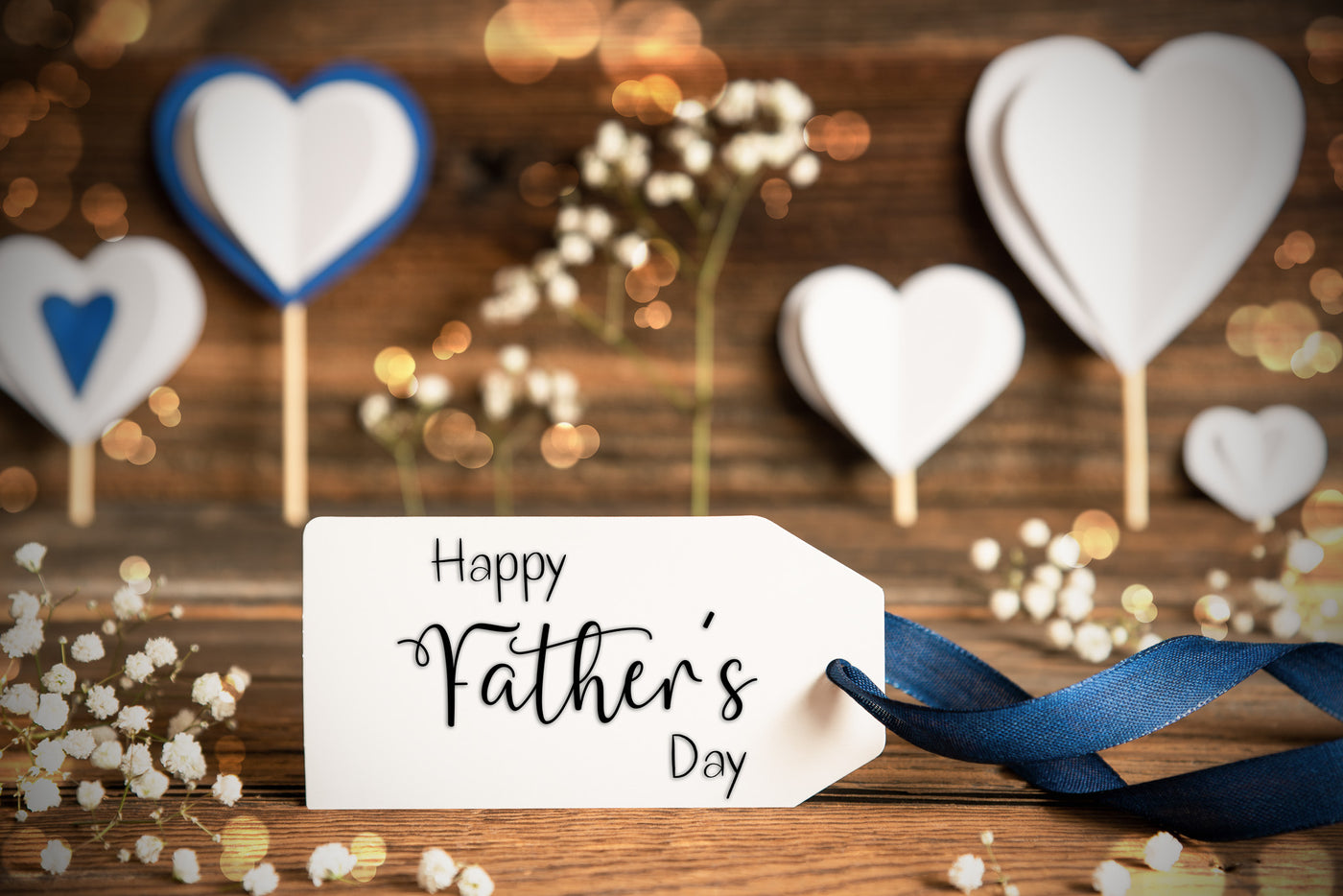 White gift tag that says Happy Father's day with blue ribbon attached on a wooden surface. White cut out hearts surround the tag.