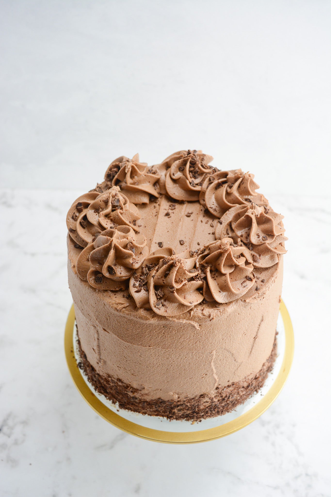 White marble background with clear cake platter with golden edge. On top sits a light brown tall cake with chocolate shavings at the base and swirls of frosting on top and chocolate shavings